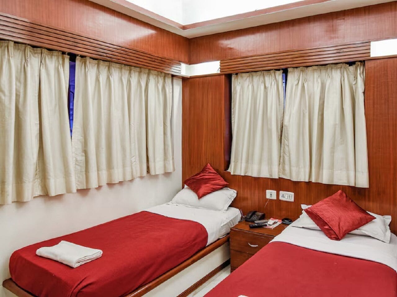 Una habitación con dos camas individuales, cada una con colcha roja y almohadas blancas. Tiene un cabecero de madera, una mesita de noche con lámpara y una ventana con cortinas.