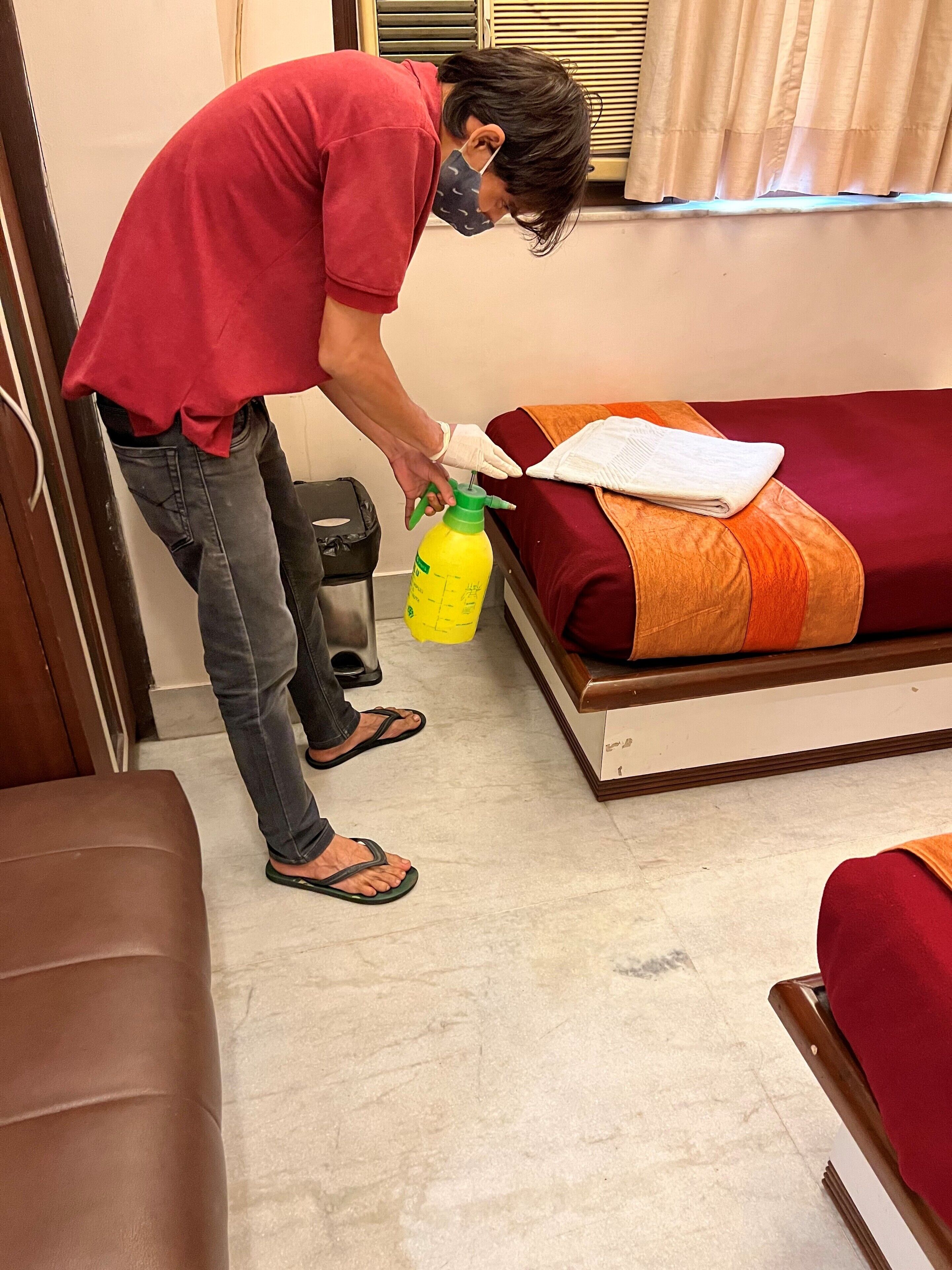 A person in a red shirt and jeans preparing to use a yellow spray bottle in a room with a bed, a wooden bench, and a trash bin.