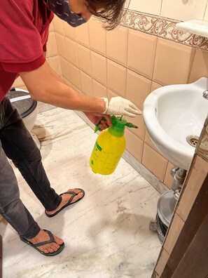 A person holding a yellow bottle with a green nozzle, standing in a bathroom with beige tiles and a white sink. - Hotel Aston  (Kolkata)