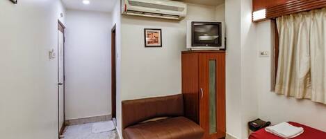 A compact hotel room with a single bed, a sofa, an air conditioning unit, a television, and a wardrobe.