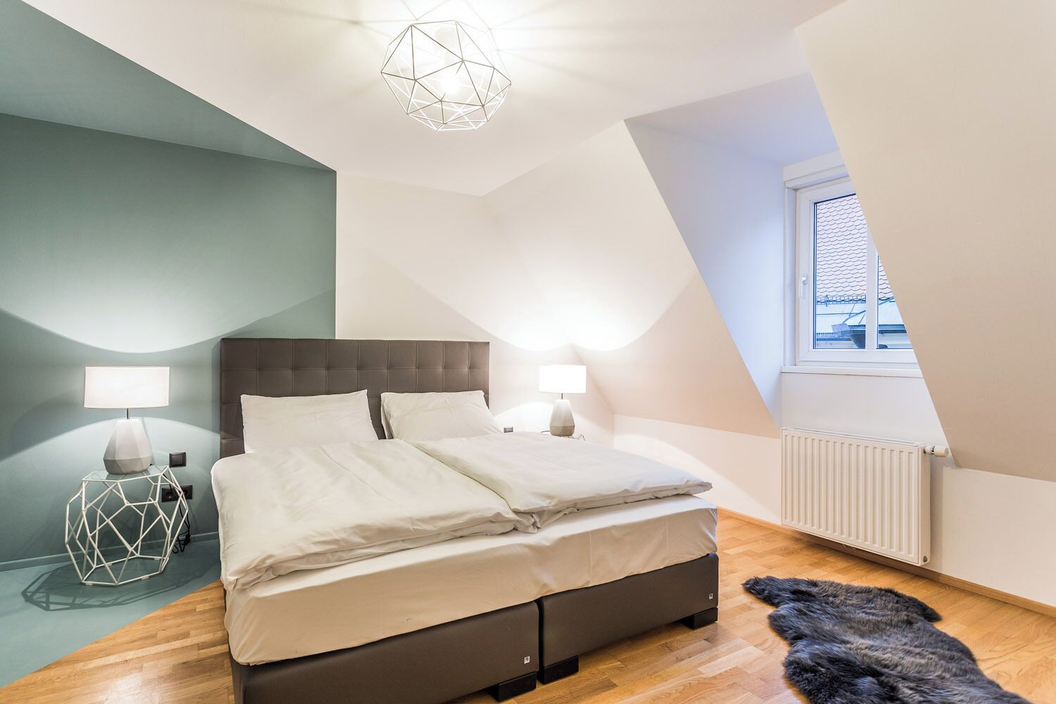 A bedroom with a bed, two bedside lamps, a geometric nightstand, a fur rug, and a window with a view.