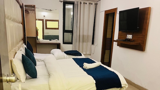 A neatly arranged bedroom with a bed, bedside tables, a TV mounted on the wall, and a green sofa. - My Home Hotels (New Delhi)