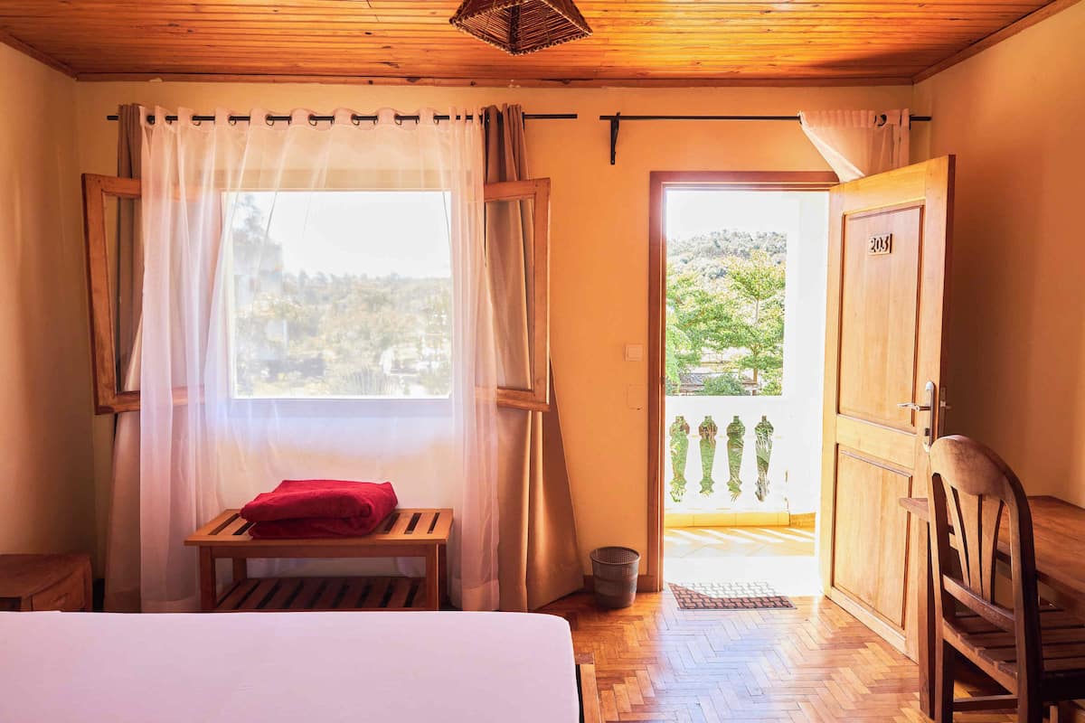 A bedroom with a bed, a wooden bench, a red towel, a wooden door, and a view of greenery outside.