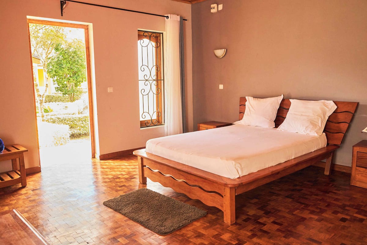 A bedroom with a wooden bed, a nightstand, a bedside table, a window with curtains, and a view of the outdoors.