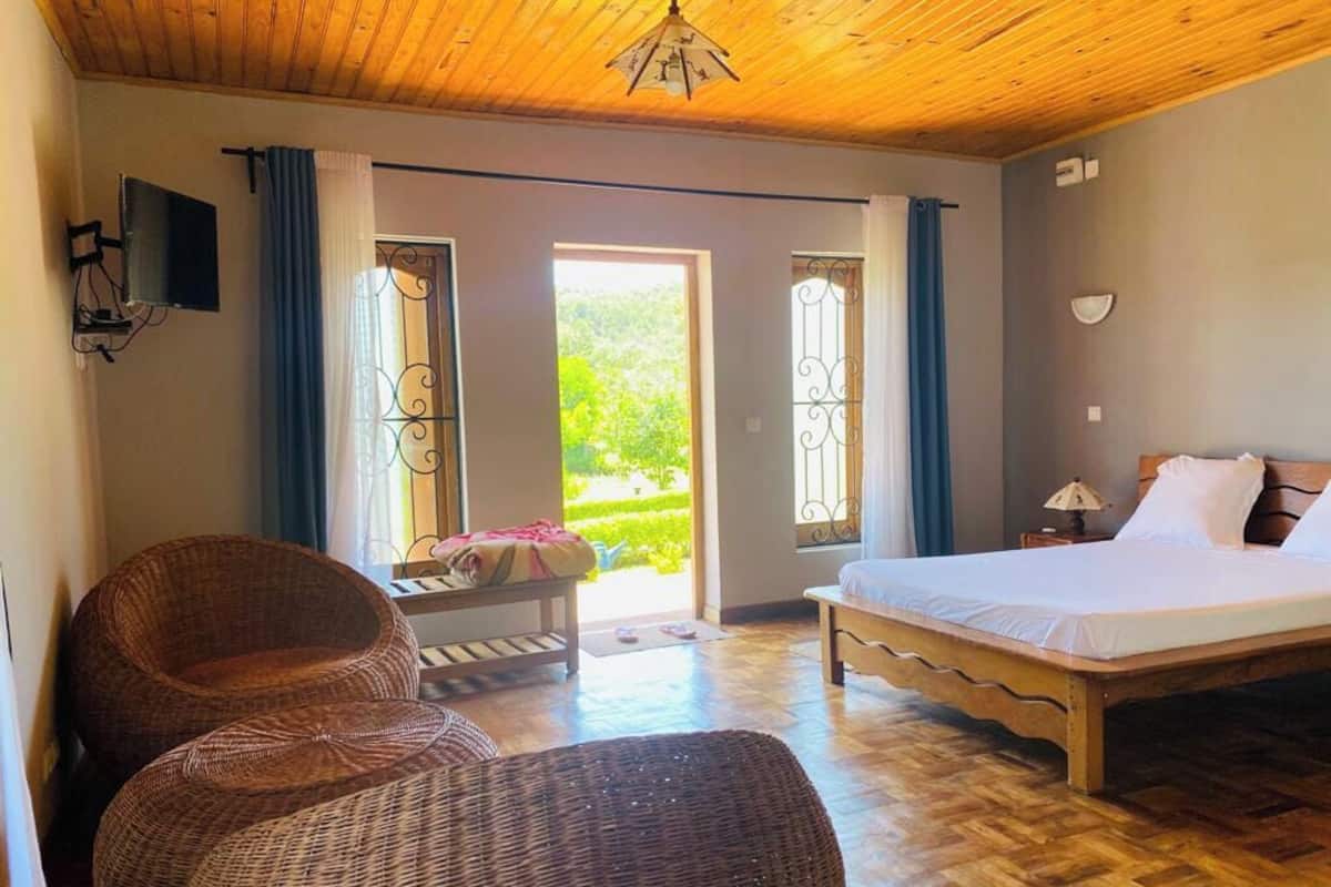 A bedroom with a bed, wicker chairs, a small table, and a view of greenery through the door.