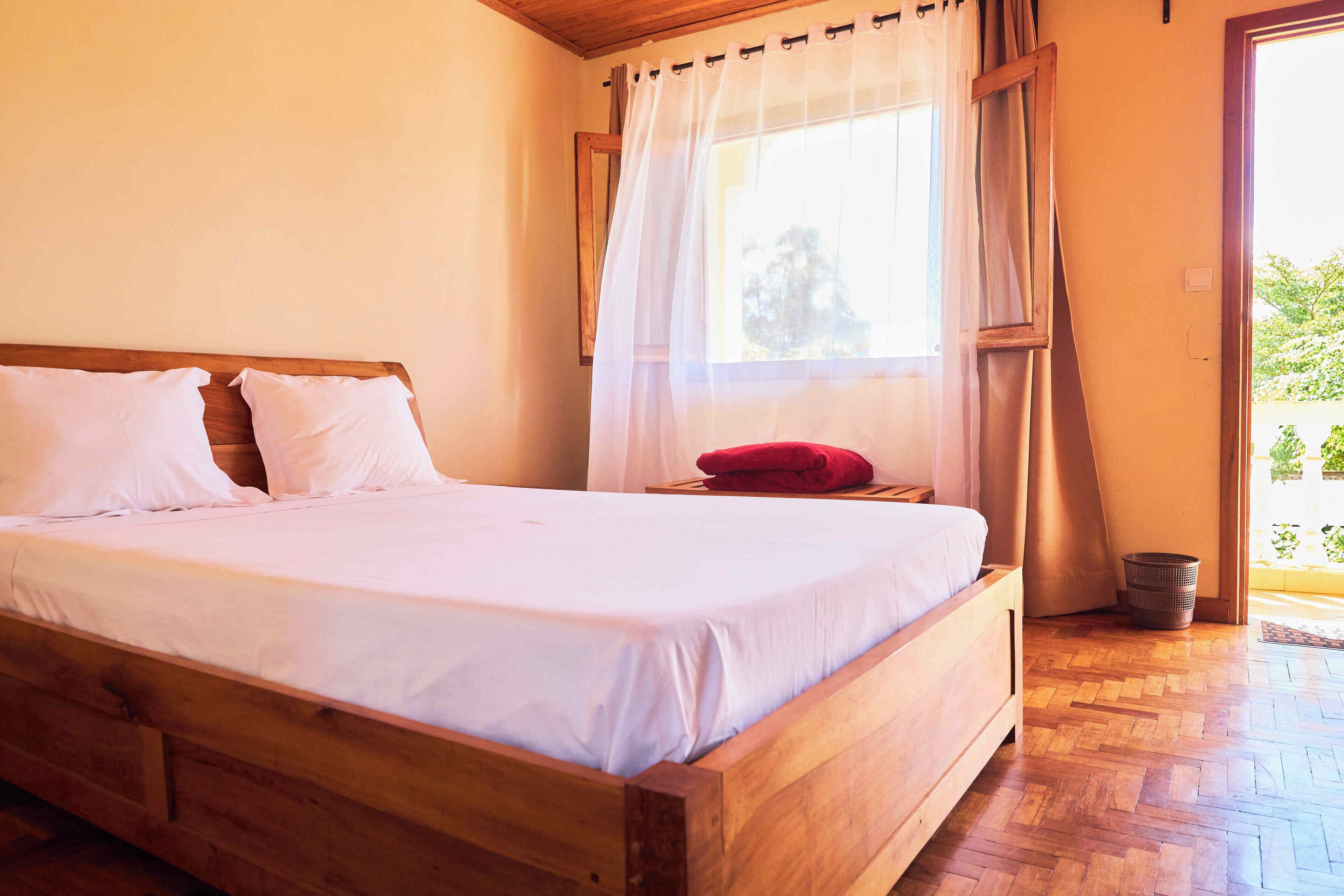 A wooden bed with white bedding in a room with a wooden floor and a window with sheer curtains.