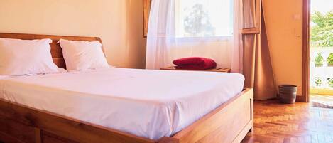 A wooden bed with white bedding in a room with a wooden floor and a window with sheer curtains.