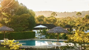 Un área de piscina con sillones de descanso y sombrillas de paja, rodeada de follaje y con vista a un edificio al fondo.
