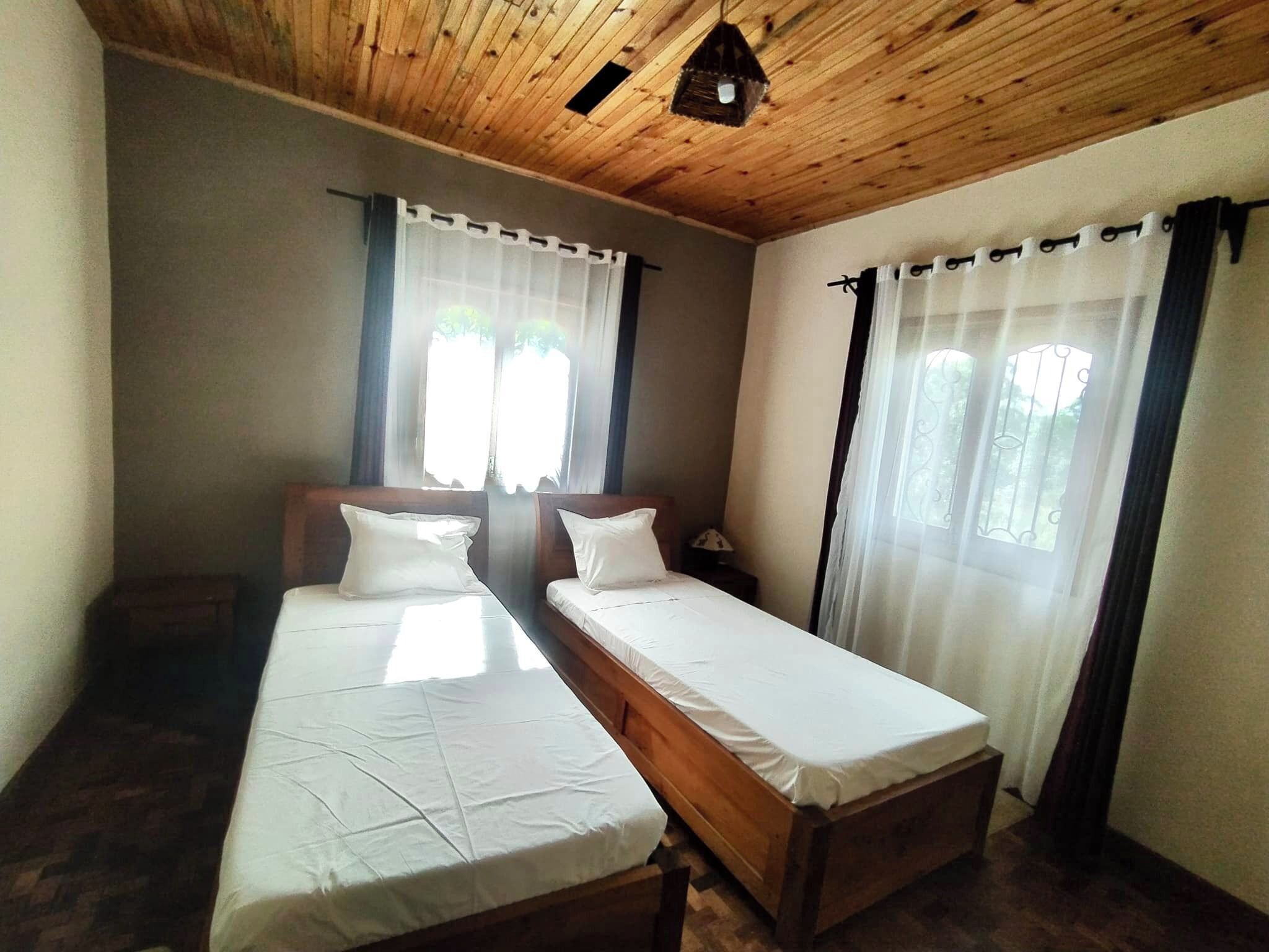 A room with two single beds, wooden ceiling, and large windows with curtains.