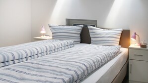 A neatly made bed with striped bedding, a bedside table with a bed lamps, and a small drawer unit. - Apartment '4, 85qm' with Shared Pool, Private Terrace and Wi-Fi (Wasserburg (Bodensee))