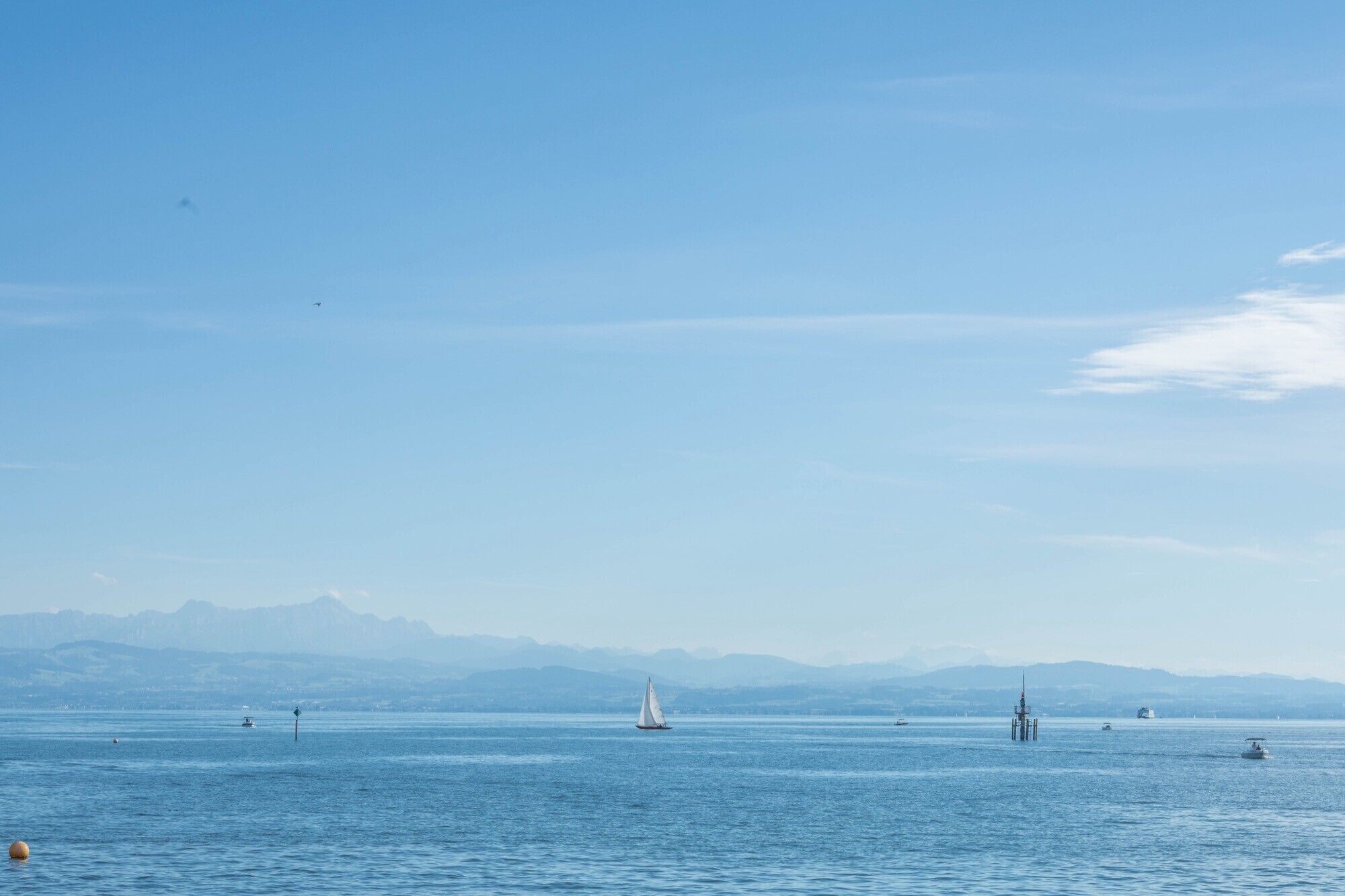 A calm body of water with a sailboat, distant mountains, and a buoy.