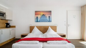 A hotel room with a bed, a wooden headboard, a painting of a sunset over a pier, a white door, and a kitchen area with a microwave. - Apartment 'Apt Typ A1' with Shared Terrace, Shared Garden and Wi-Fi (Immenstaad am Bodensee)