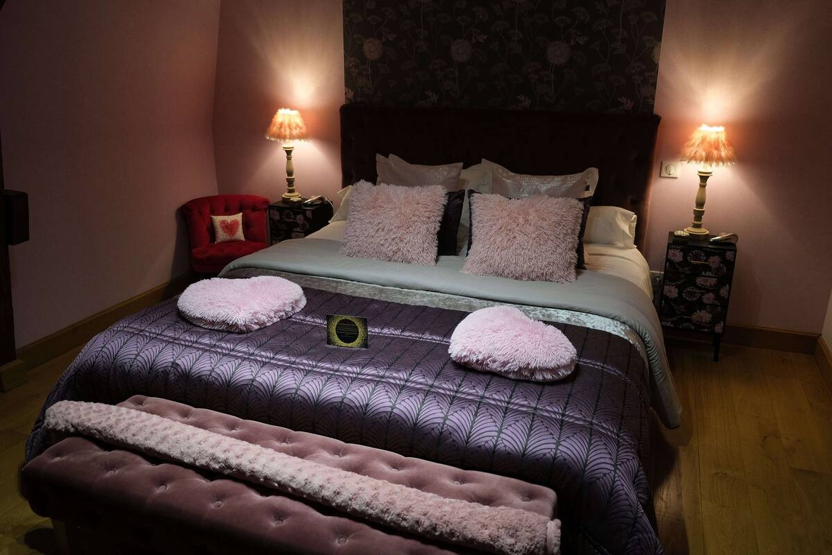 Una cama bien hecha con una colcha estampada, dos lámparas de noche, una silla roja y un banco.