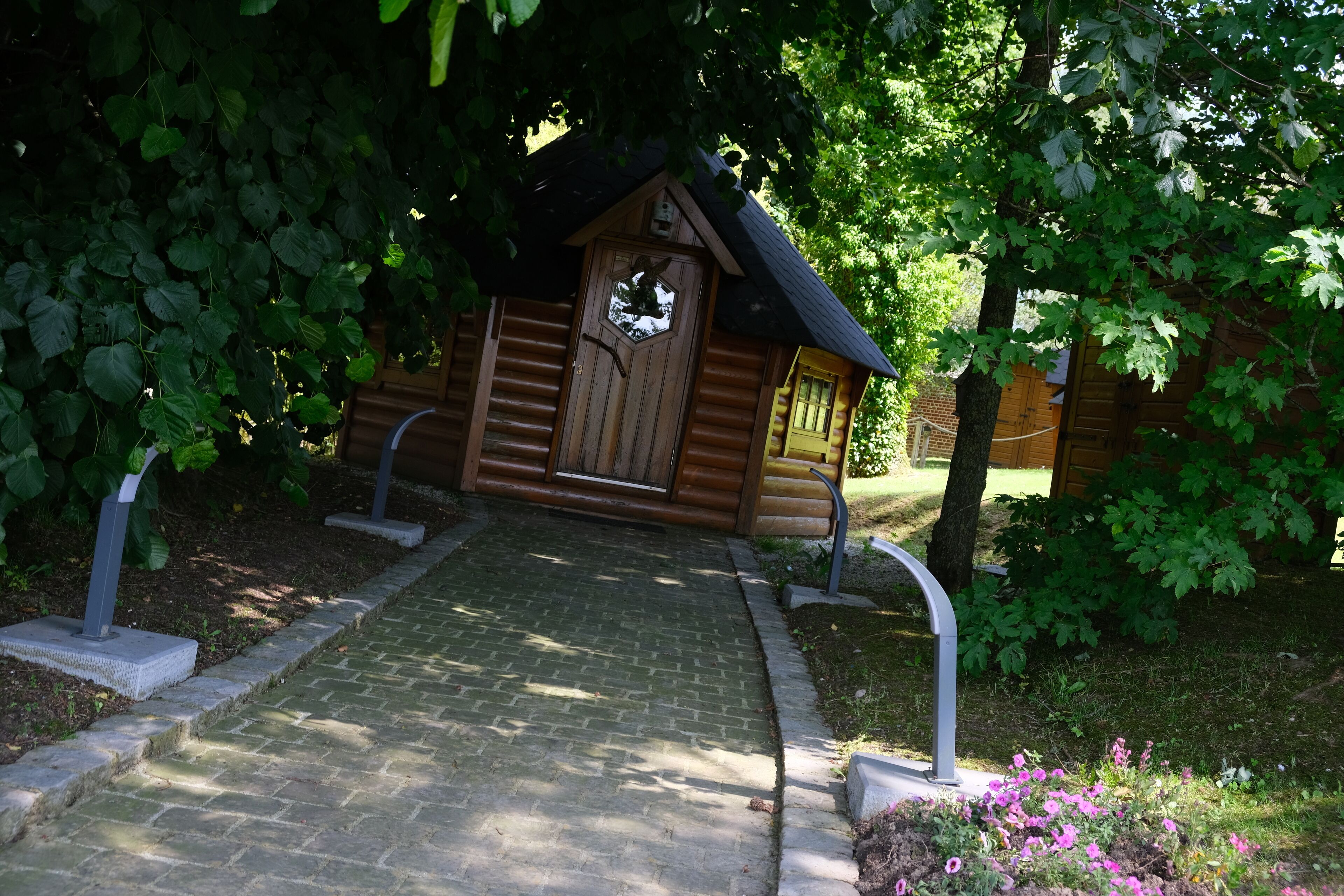 A wooden cabin with a hexagonal window and a door, surrounded by greenery and a brick pathway.