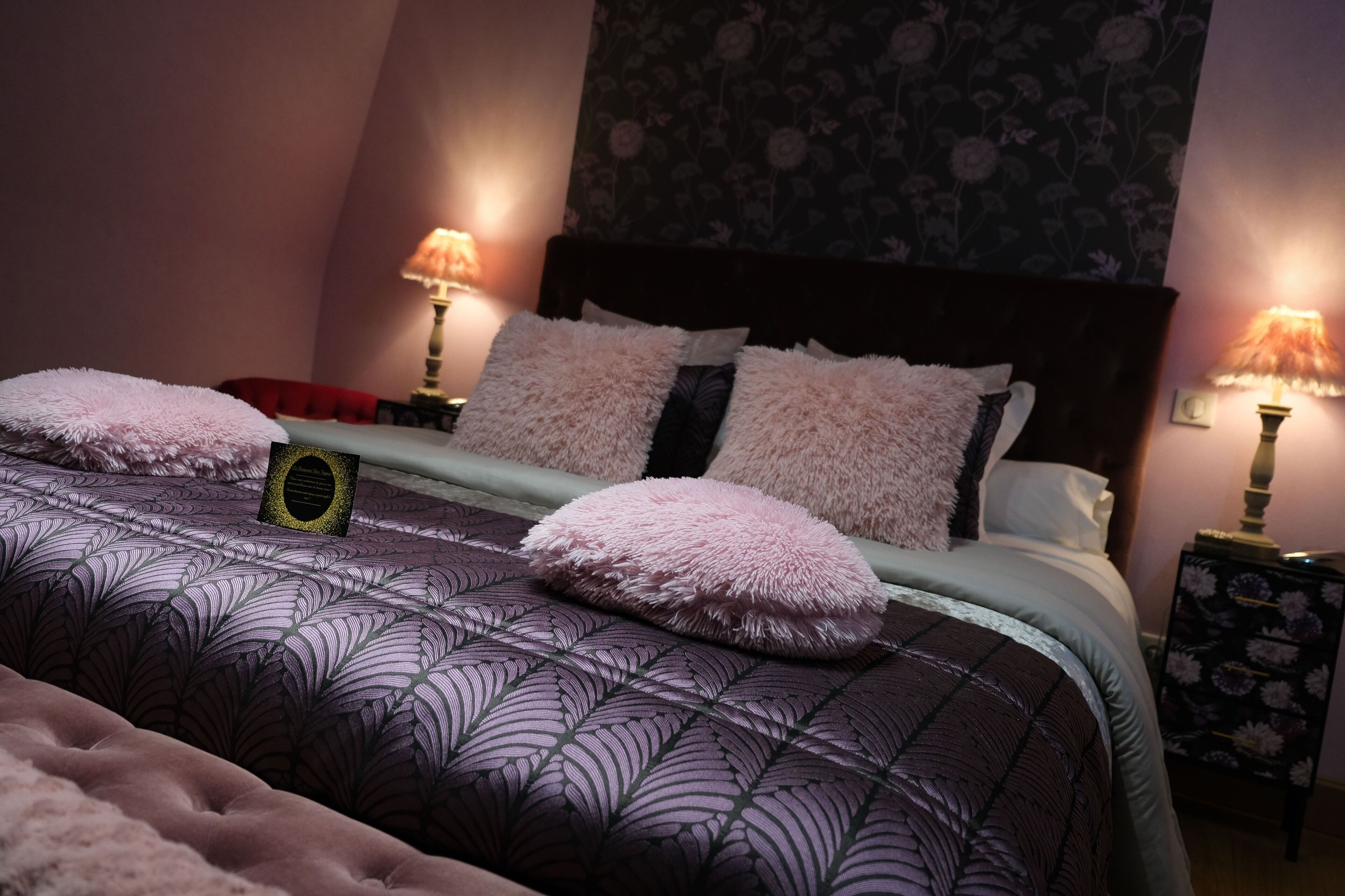 A neatly made bed with a patterned bedspread and fluffy pillows, two bedside lamps, and a floral wallpaper backdrop.