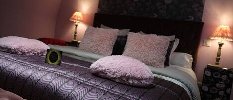 A neatly made bed with a patterned bedspread and fluffy pillows, two bedside lamps, and a floral wallpaper backdrop.