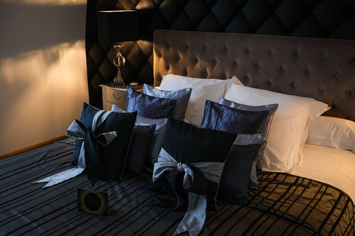 Una cama bien hecha con cabecera capitonada, una mesita de noche con lámpara, y un papel tapiz decorativo en la pared.