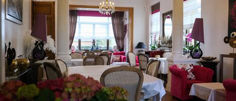 A dining area with tables and chairs, a chandelier, and a view through large windows.