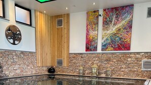 A modern indoor pool with a stone wall and wooden accents, featuring a large Buddha statue and abstract art. - Le Domaine Des Fagnes (Sains-du-Nord)