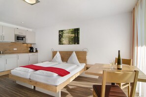 A modern hotel room with a bed, a wooden headboard, a dining table with chairs, a kitchen area, and a framed picture on the wall.