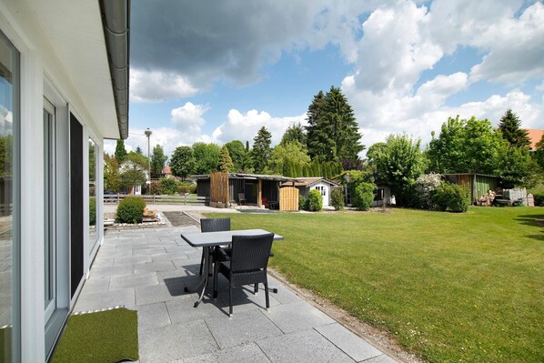 Une terrasse avec une table et des chaises, une cour gazonnée et un bâtiment doté d’un toit en pente.