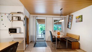 Une cuisine équipée d’un four à micro-ondes, d’un grille-pain et d’étagères, un coin repas avec un banc, et une vue sur l’extérieur par une porte.