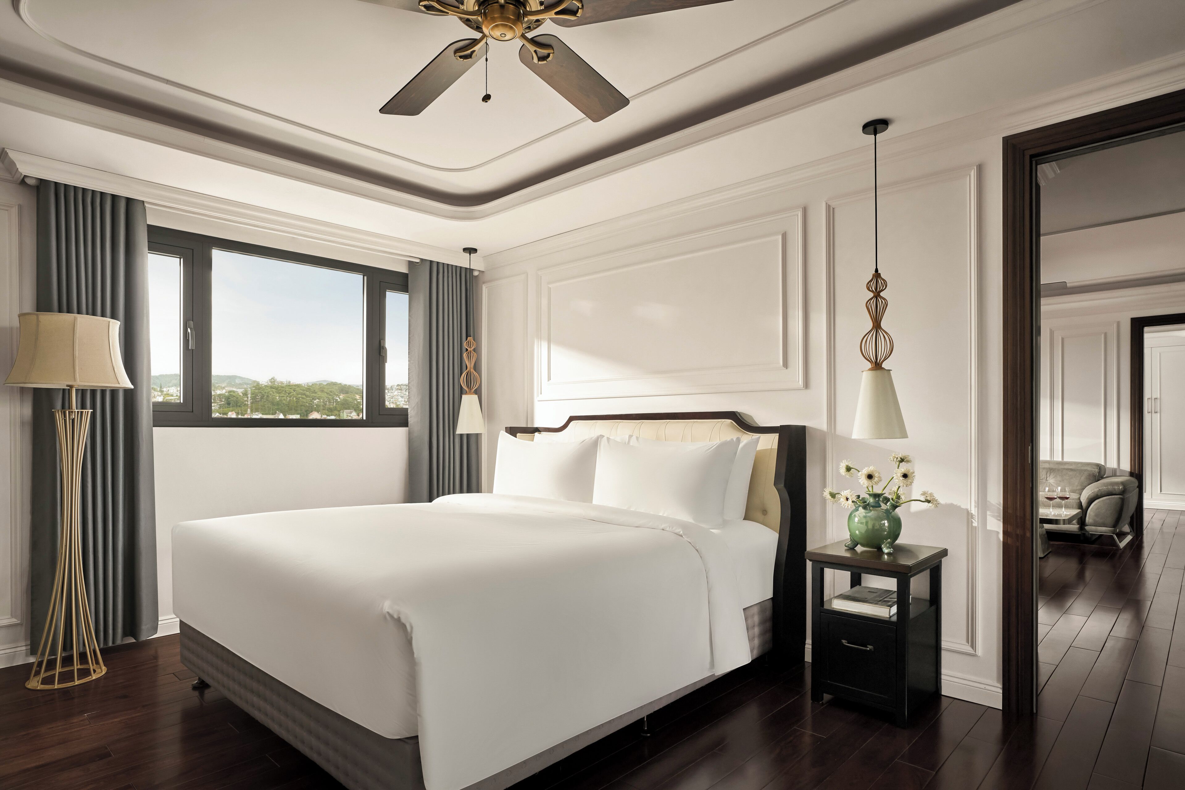 A hotel room with a large bed, a bedside table with a vase, a floor lamp, and a view of the outdoors.