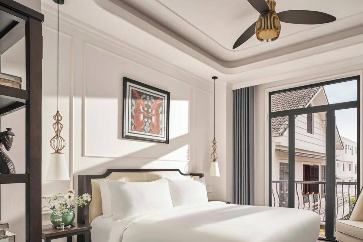 A hotel room with a bed, a chair, a desk, a window with a view, a ceiling fan, and artwork on the wall.