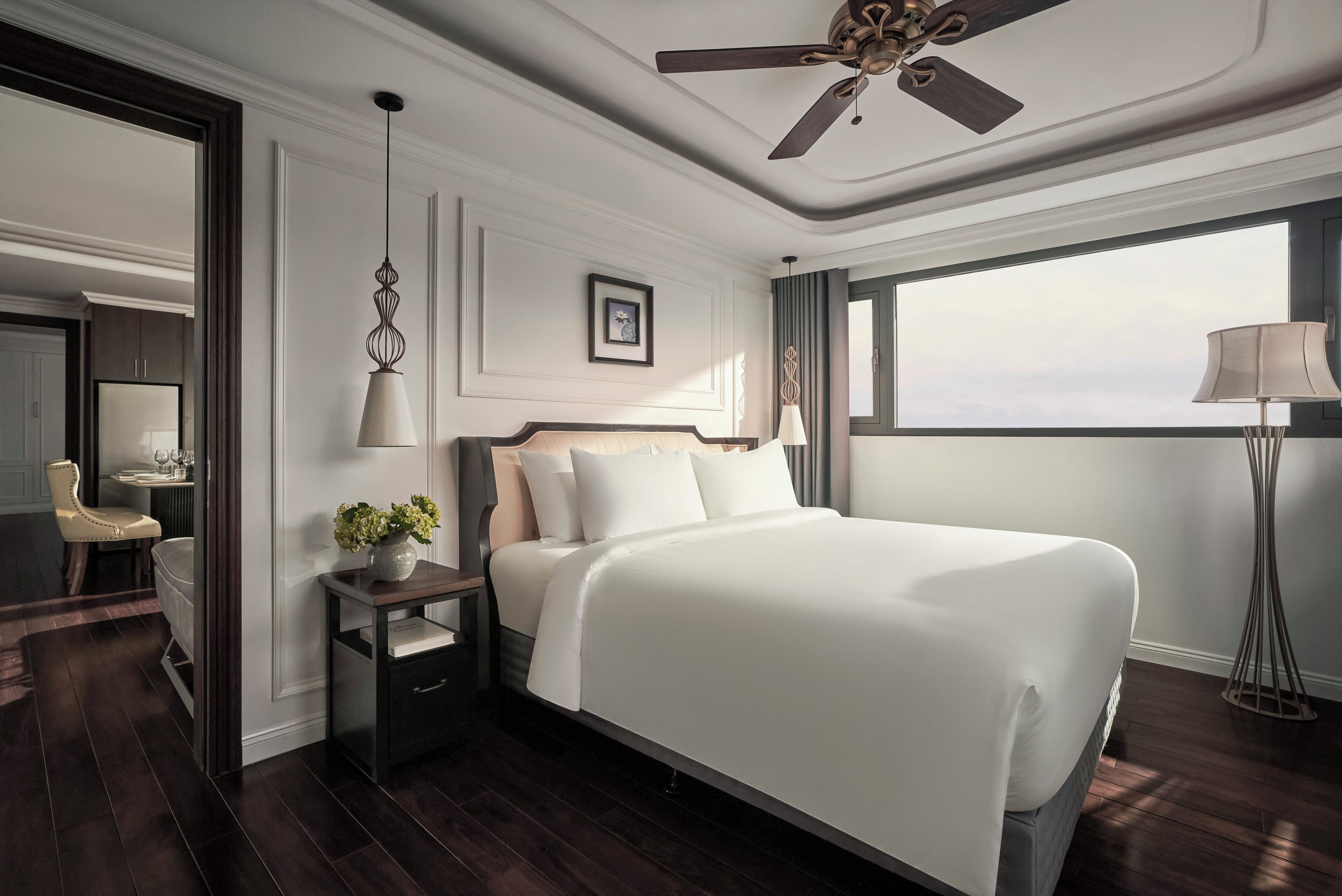 A hotel room with a large bed, a ceiling fan, a bedside table with a lamp, a nightstand, a chair, and a view of the sky through a window.