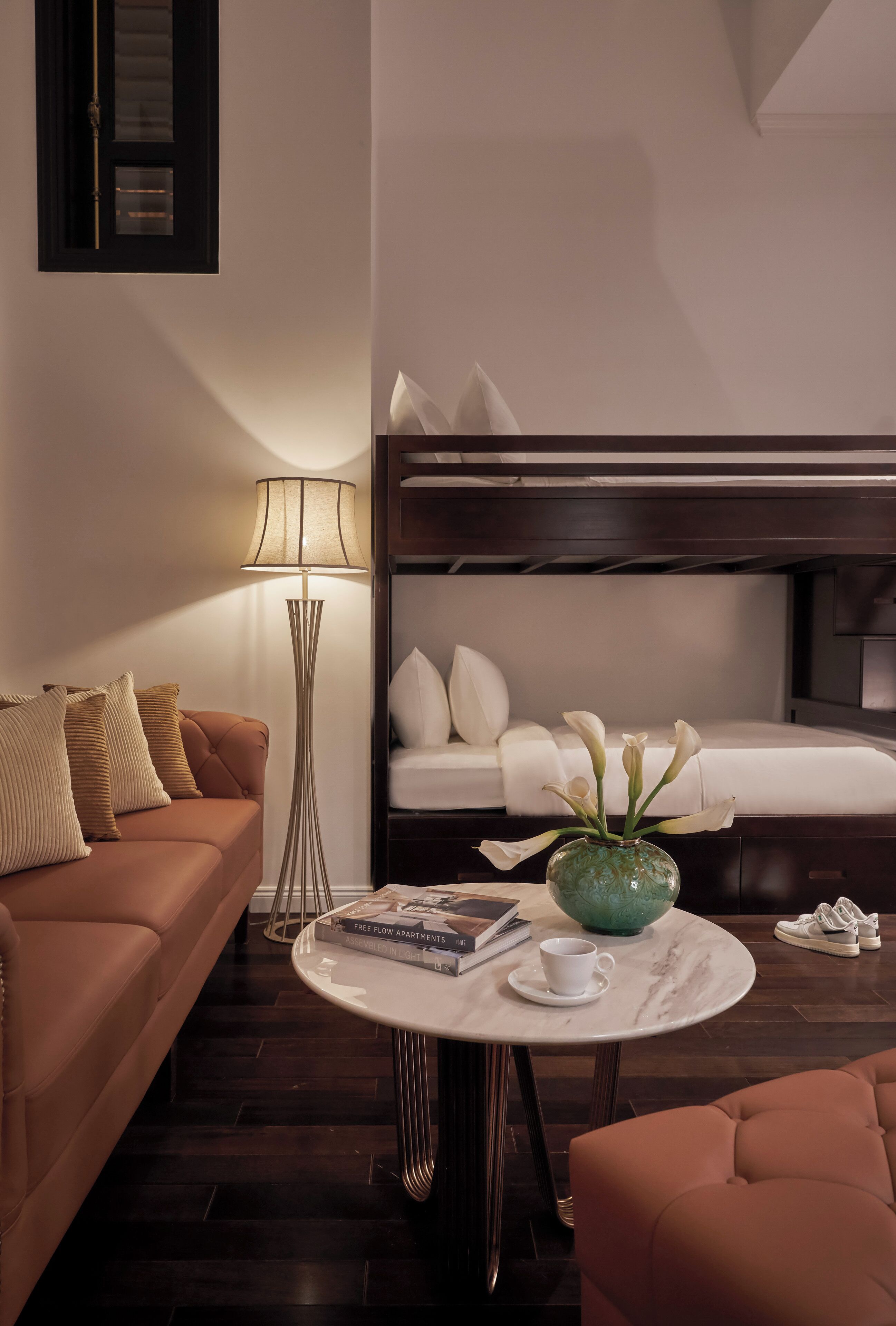Une chambre d’hôtel avec un canapé marron, une table ronde ornée d’un vase et de livres, une table de chevet avec une lampe, et un lit superposé avec une literie blanche.