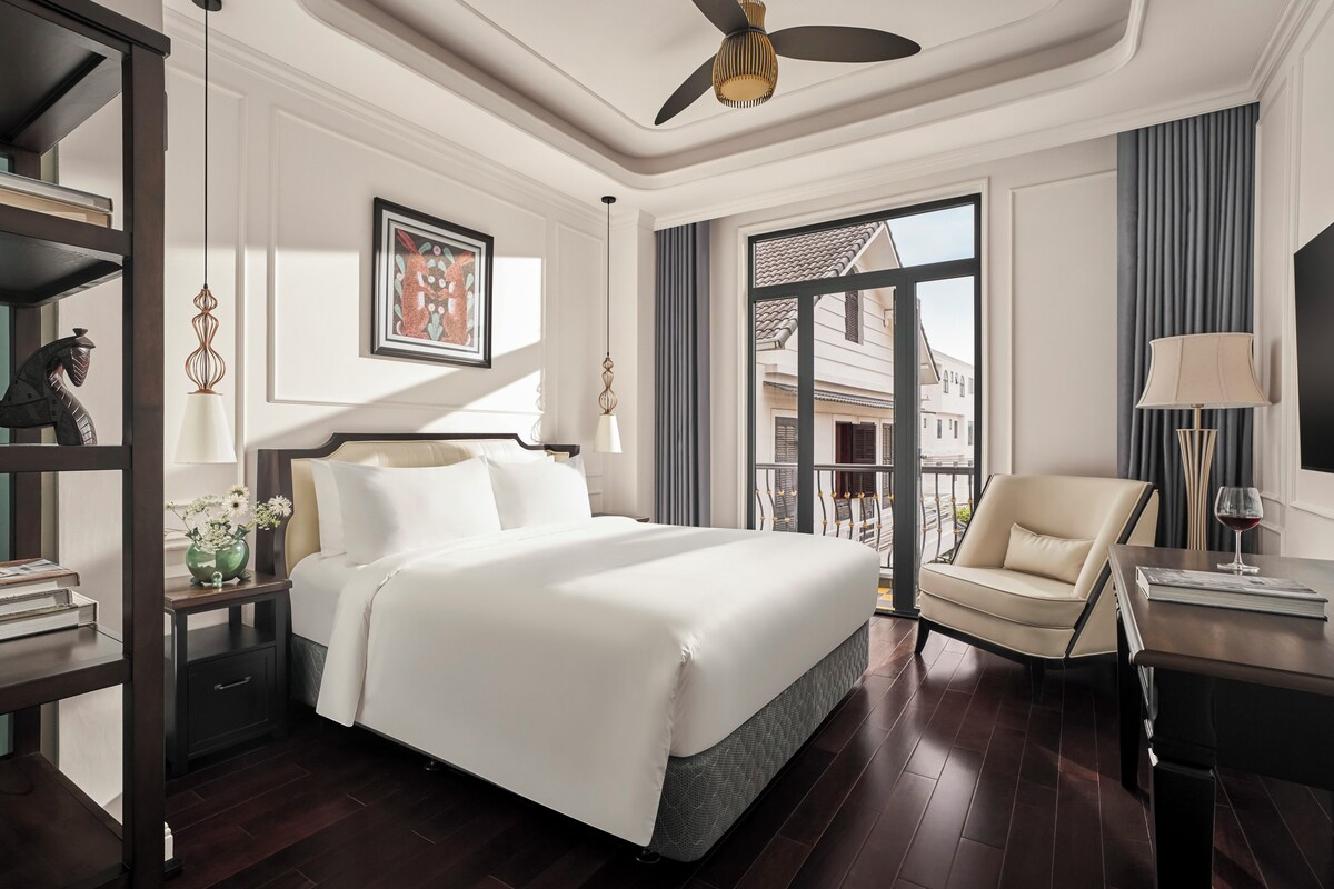 A hotel room with a large bed, a chair, a desk with a lamp, a bookshelf, and a window with a view of buildings.