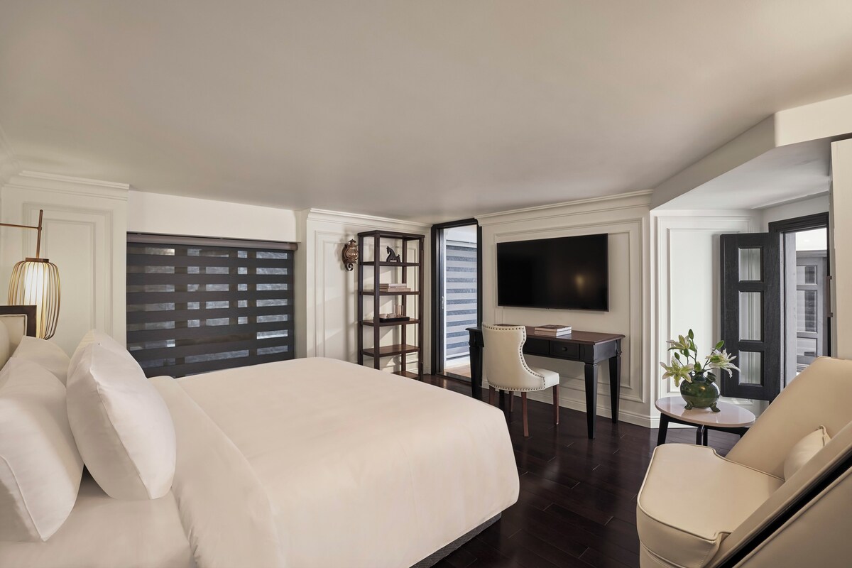 A hotel room with a large bed, a desk, a chair, a television, and a small table with a plant.