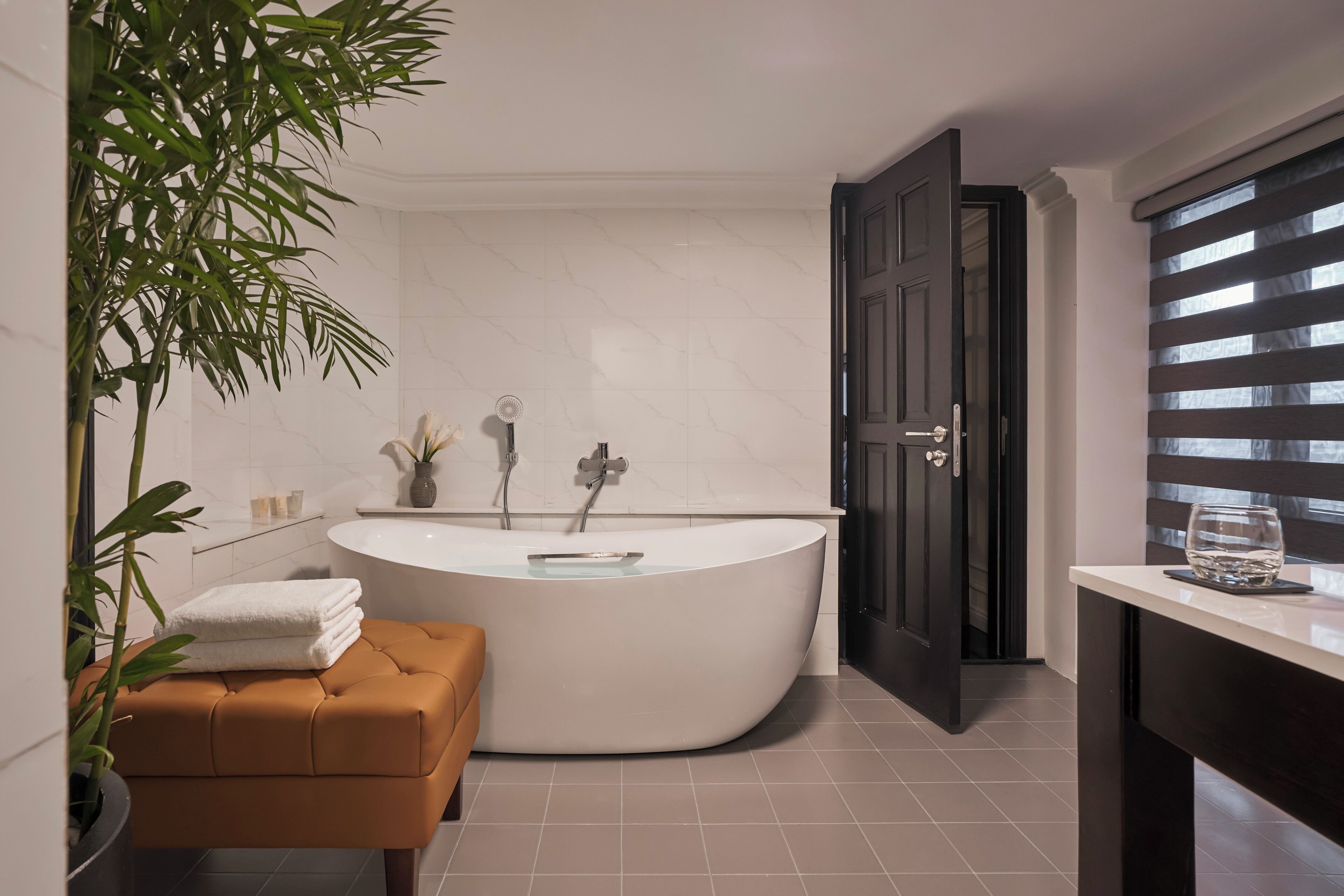 Een moderne badkamer met een vrijstaand bad, een potplant, een bankje en een deur naar een andere ruimte.