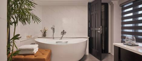 A modern bathroom with a freestanding bathtub, a potted plant, a bench, and a door leading to another room.