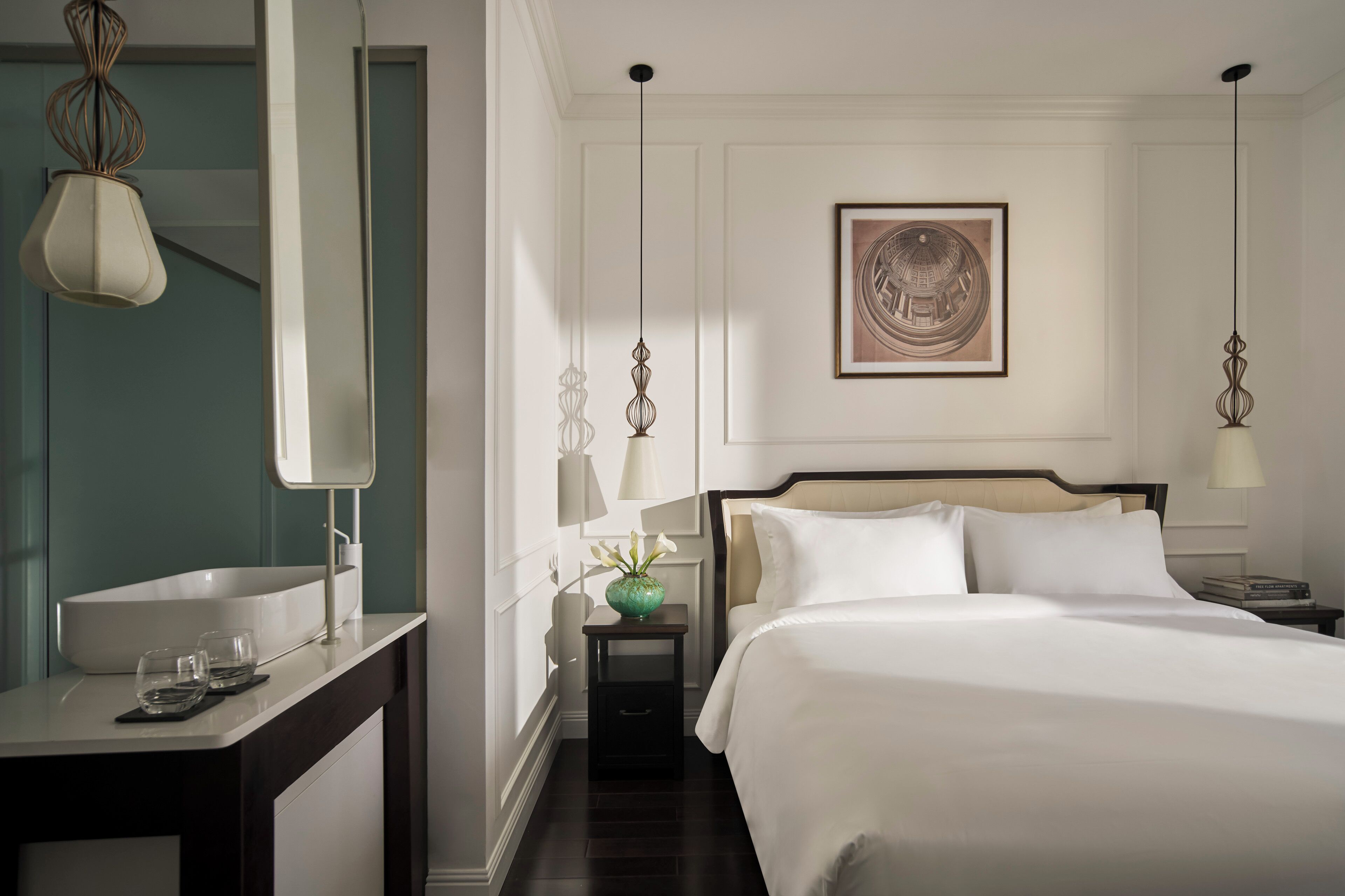 Habitación de hotel con cama, mesitas de noche, un espejo, lavabo y un florero con flores.