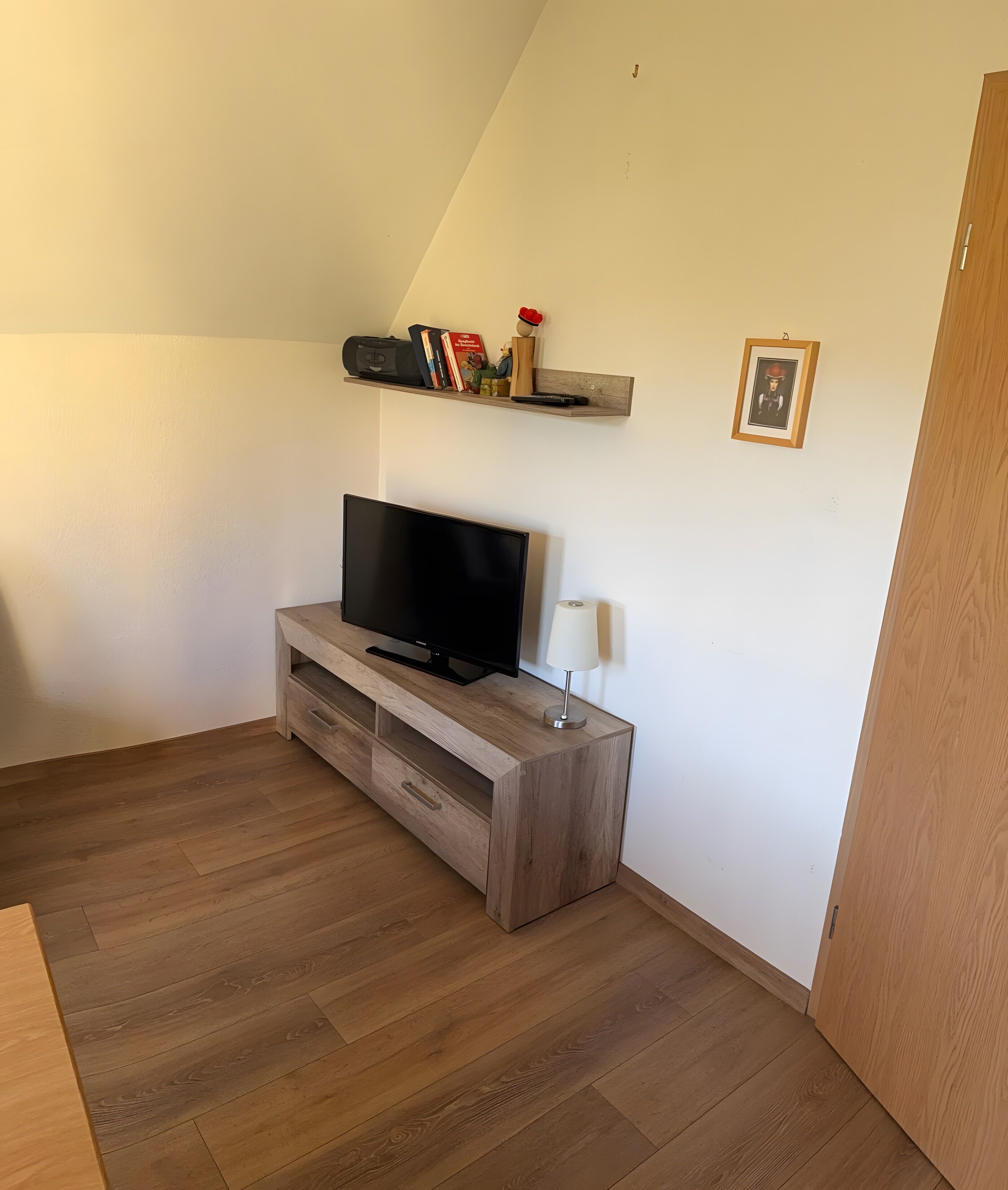 A room with a flat-screen TV on a wooden TV stand, a small shelf with books and decorative items, a white lamp, and a wooden door.