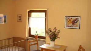 Une pièce comprenant un berceau, une chaise en bois, une table avec un pot de plante, une fenêtre avec des rideaux et deux tableaux encadrés accrochés au mur.