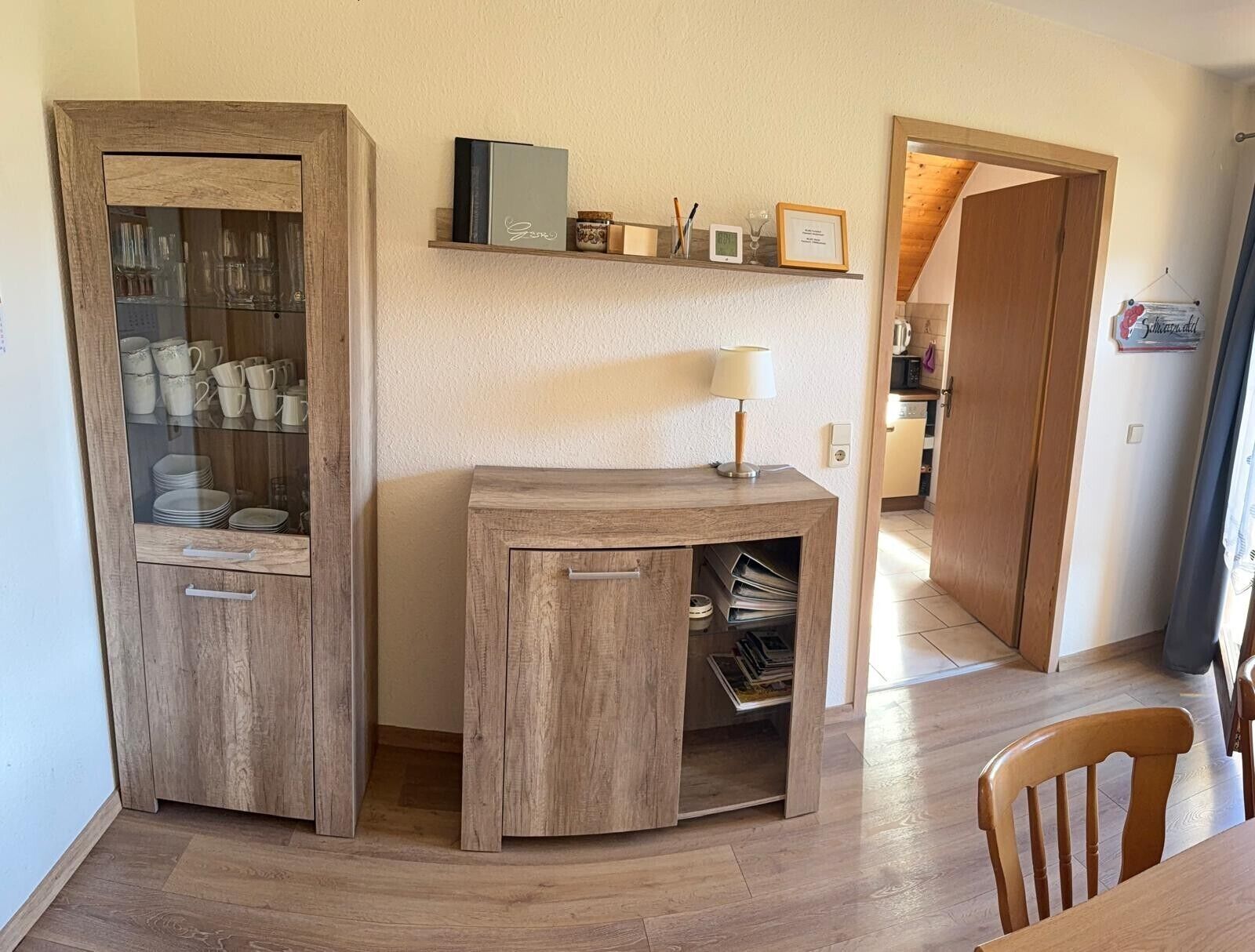 Una habitación con un armario de madera con puertas de cristal, un escritorio con lámpara y una silla.