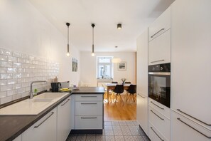 Una cocina moderna con armarios blancos, encimera negra y salpicadero de azulejos. Se ve un área de comedor con mesa y sillas al fondo.