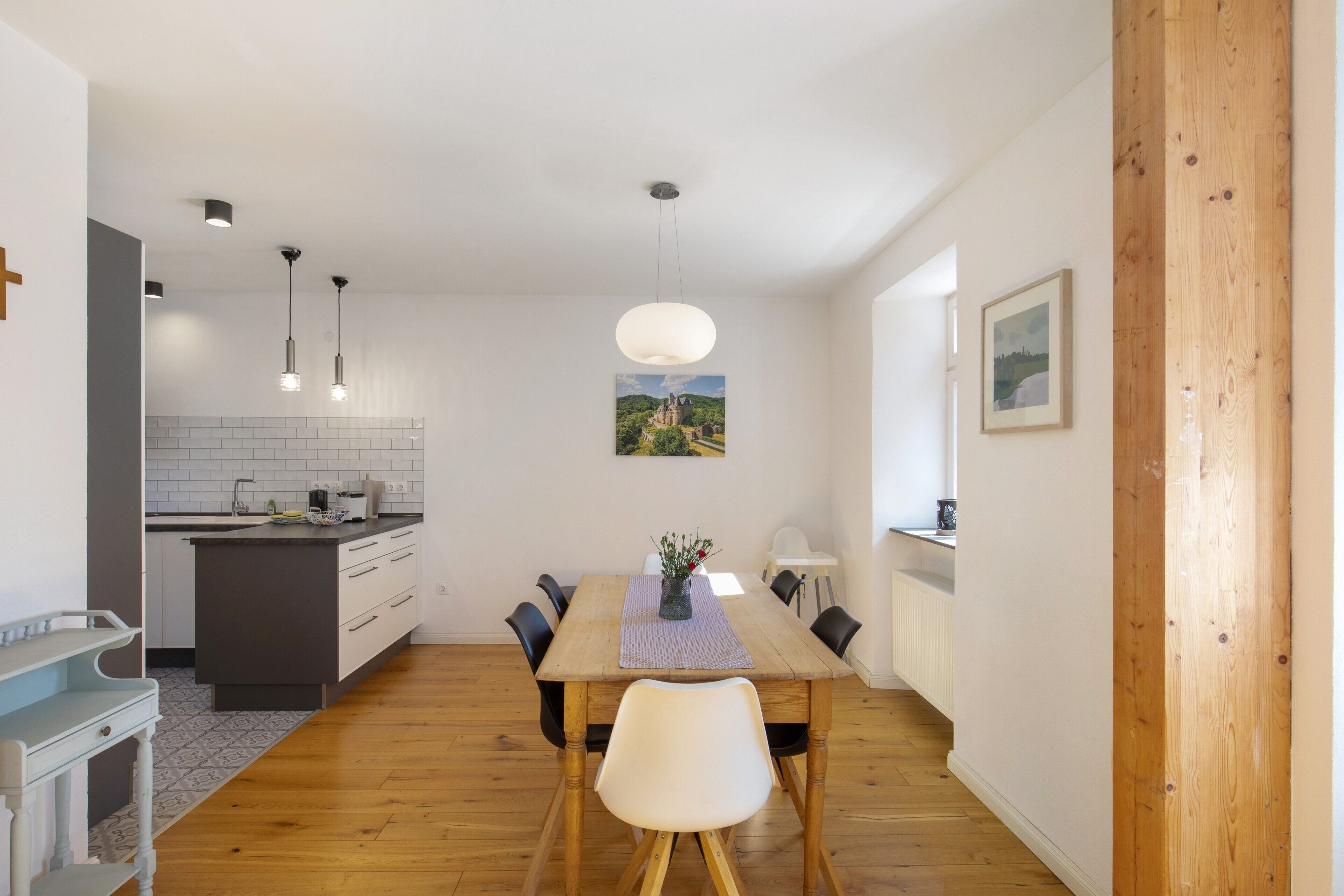 A modern kitchen with a wooden dining table, pendant lights, and a view of a landscape painting.