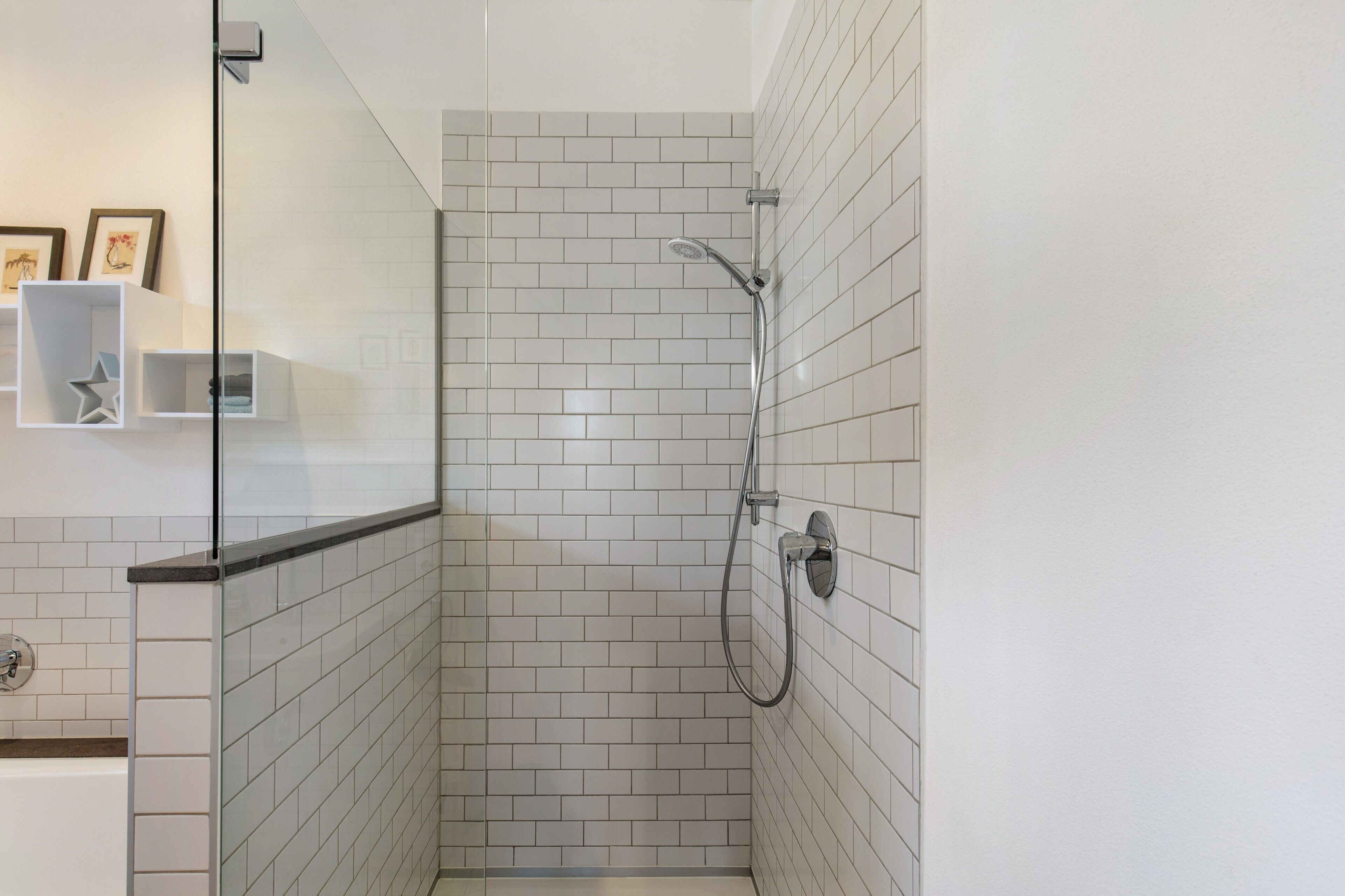 Eine Eckdusche mit weiß gefliesten Wänden, einer Glasabtrennung und einer integrierten Ablage mit gerahmten Bildern.