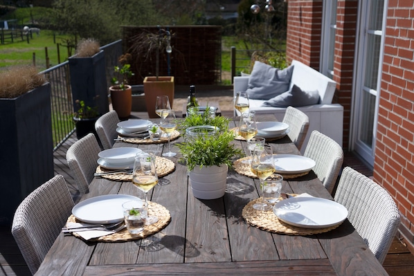 An outdoor dining area with a wooden table set for six, surrounded by wicker chairs, with a potted plant in the center.