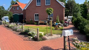 Een gebouw van rode baksteen met een schuin dak, een groene tuinhuisje, en een verhard gebied met een bankje en een brievenbus.