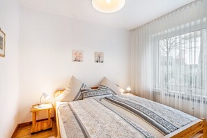 Une chambre à coucher comprenant un lit, des tables de chevet, une fenêtre avec des rideaux légers et deux tableaux accrochés au mur.
