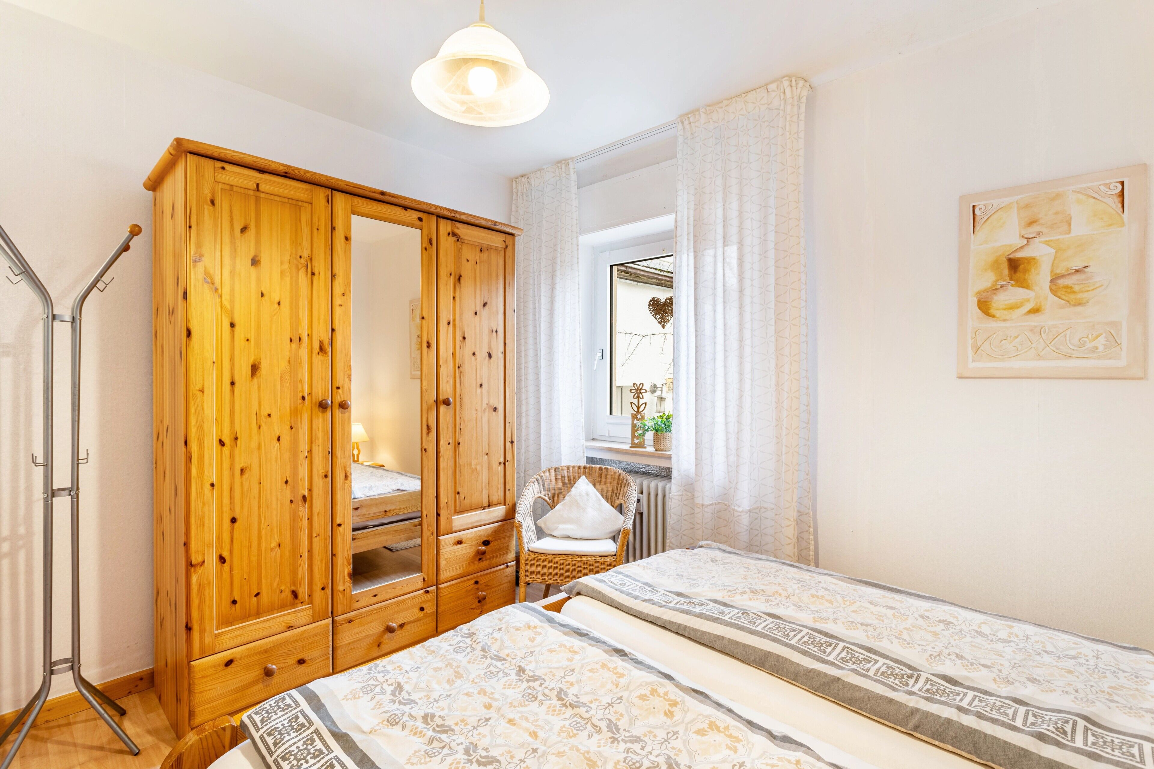 A bedroom with a wooden wardrobe, a wicker chair, a bed with patterned bedding, and a painting on the wall.