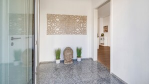 Un pasillo con una puerta de cristal, un busto de piedra, dos plantas en macetas y un adorno en la pared.