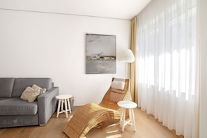 En moderne stue med en grå sofa, en fletstol, et lille hvidt bord og lette gardiner.