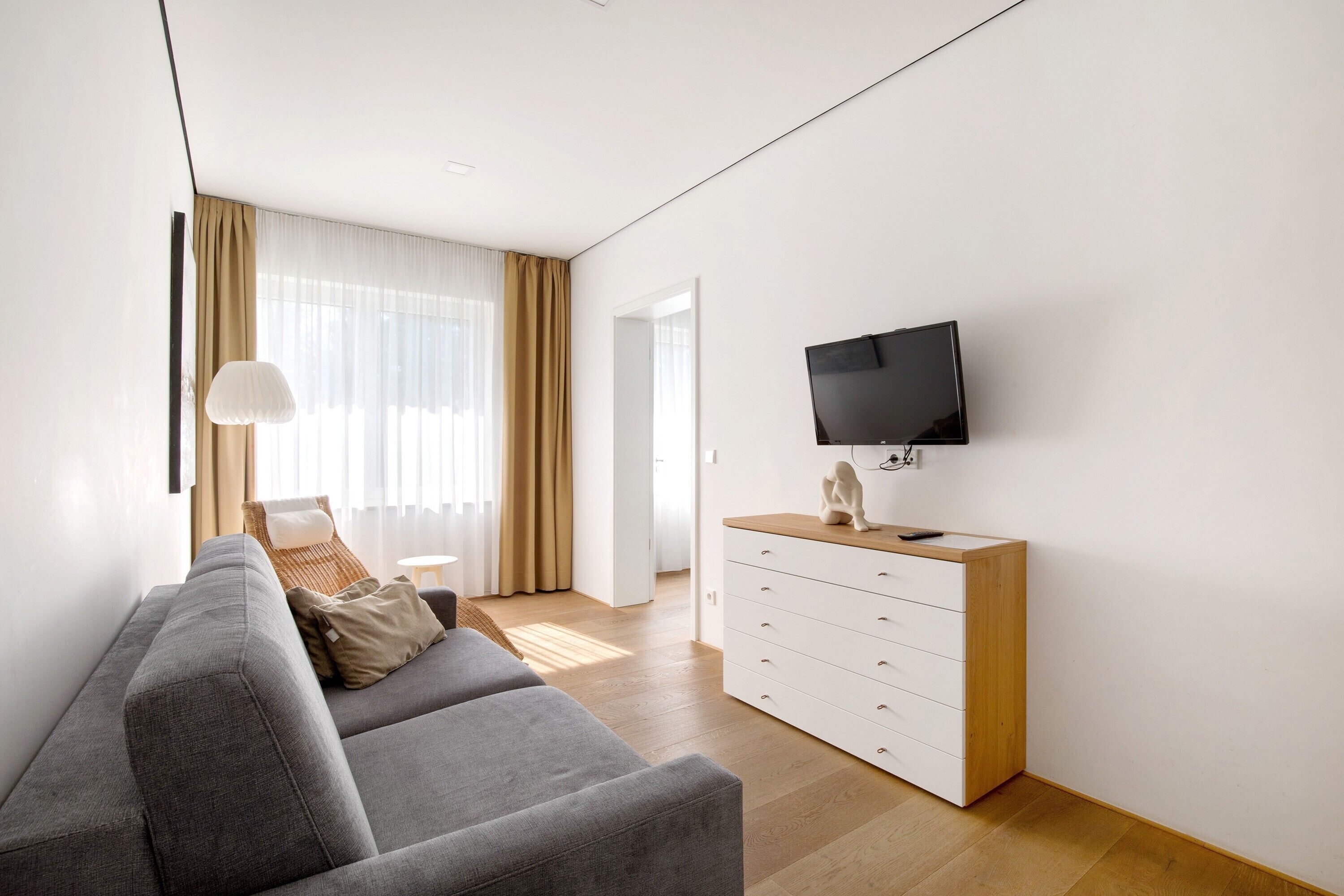 Een moderne woonkamer met een grijze bank, een houten dressoir, een flatscreen televisie en een groot raam met gordijnen.