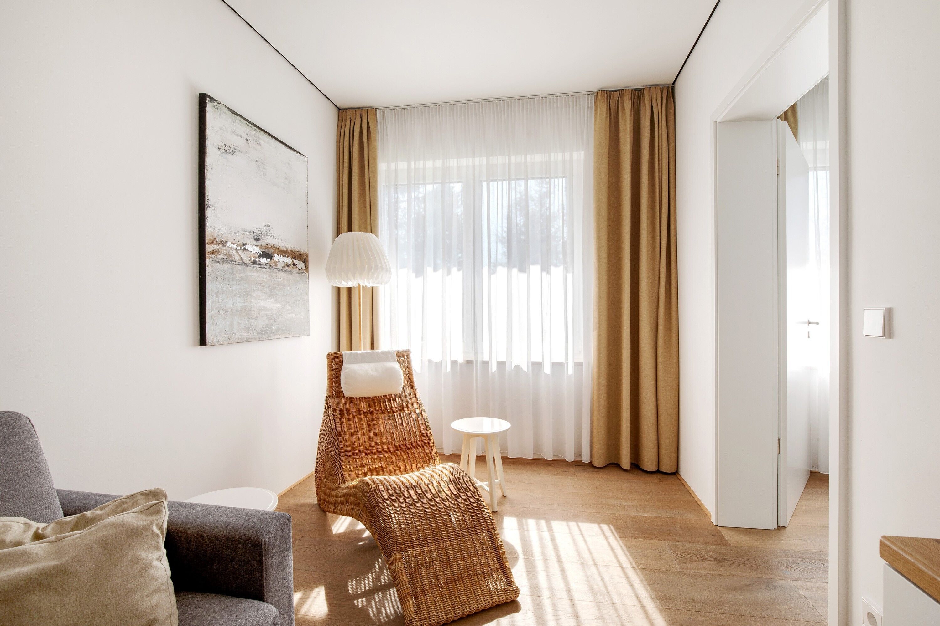Ein Zimmer mit einer Couch, einem Korbstuhl, einem kleinen Tisch, einer Lampe und einem Bild an der Wand.