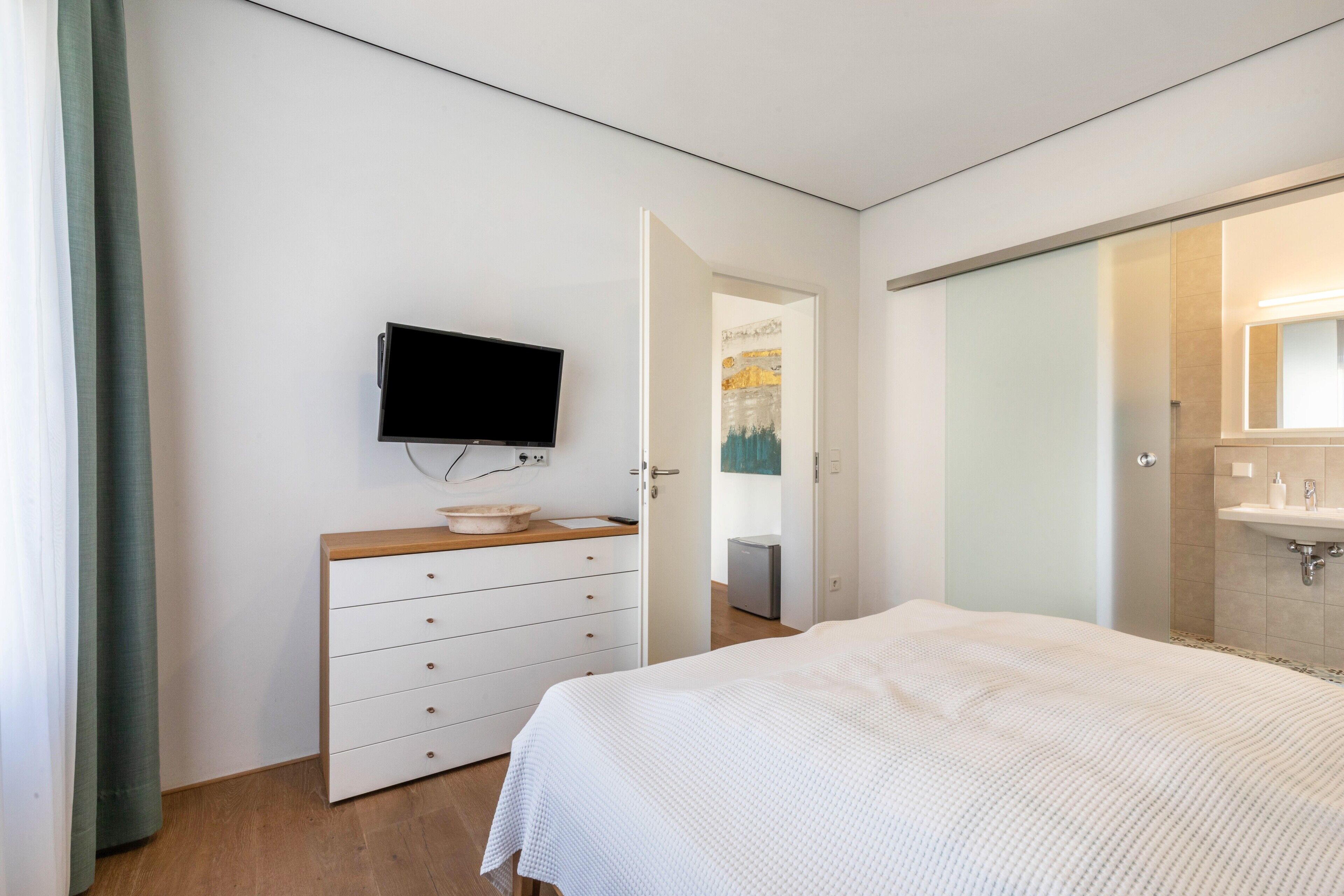Ein Schlafzimmer mit Bett, Kommode, Fernseher und einem Badezimmer mit Waschbecken und Spiegel.